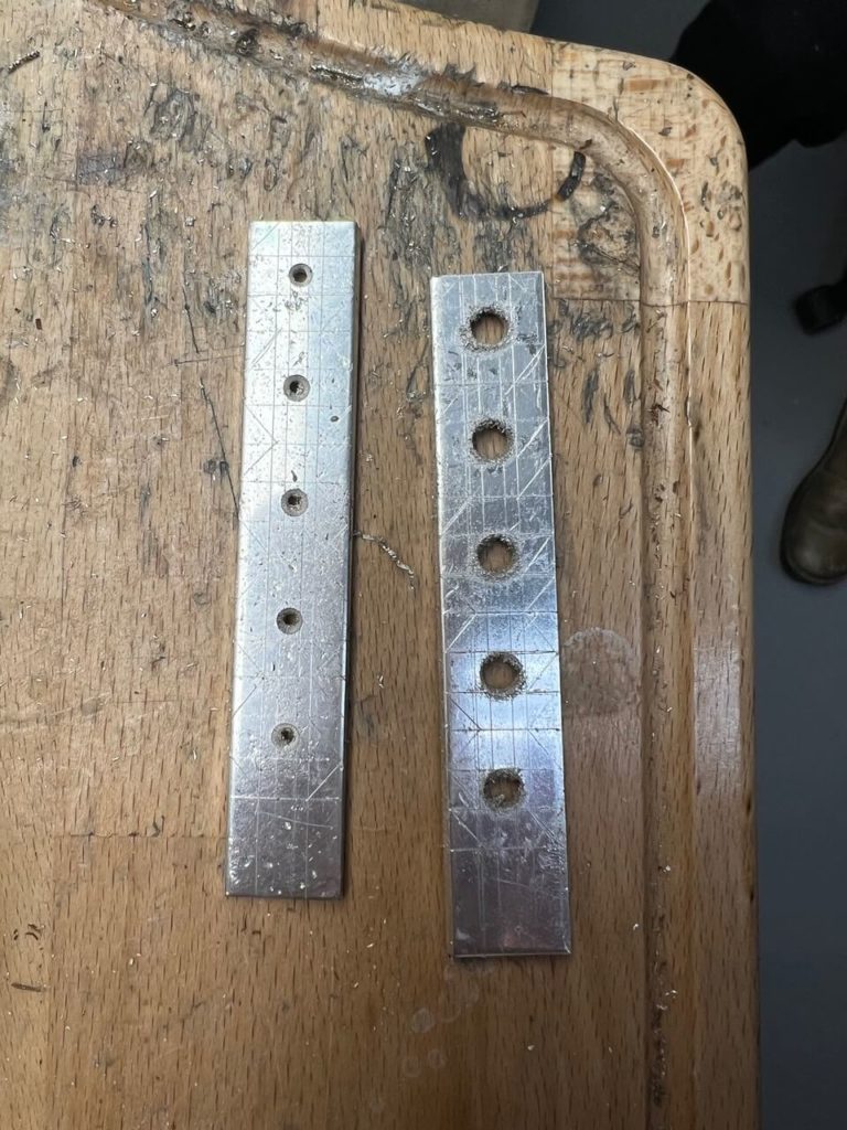 Zwei bearbeitete Metallstreifen mit Bohrungen auf einer hölzernen Werkbank in der Goldschmiedewerkstatt von Goldwerk in Berlin-Lankwitz.