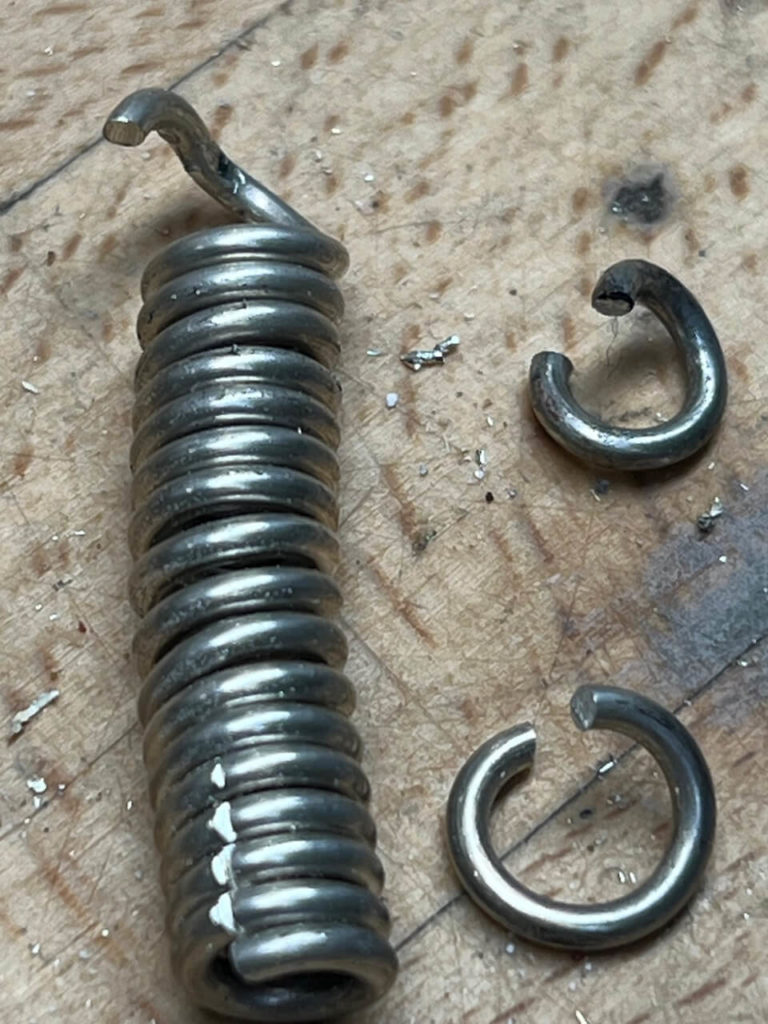 Nahaufnahme einer silberfarbenen Spiralfeder und zwei kleiner Ringe auf einer Werkbank, als Teil einer professionellen Schmuckreparatur bei Goldwerk.