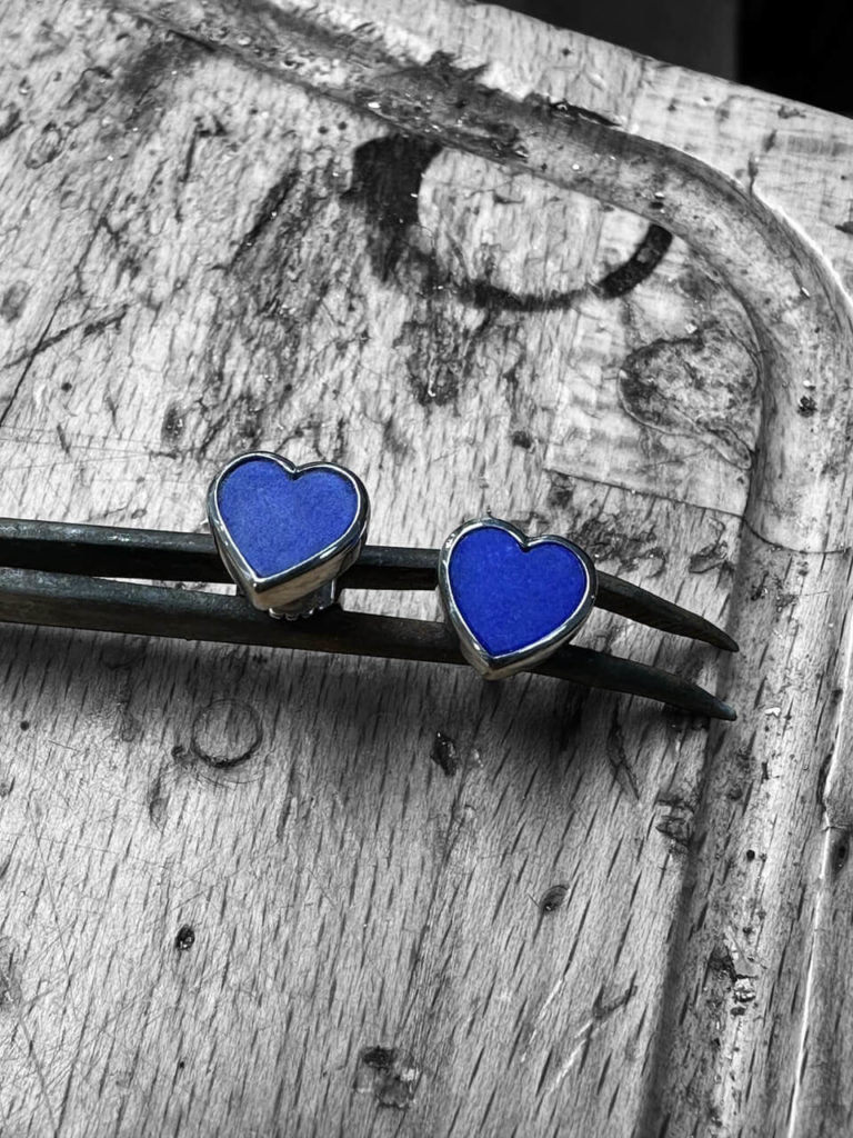 Zwei herzförmige Ohrstecker mit blauem Stein in einer Metallfassung, gehalten von einer Pinzette auf einer hölzernen Werkbank in Berlin-Lankwitz.