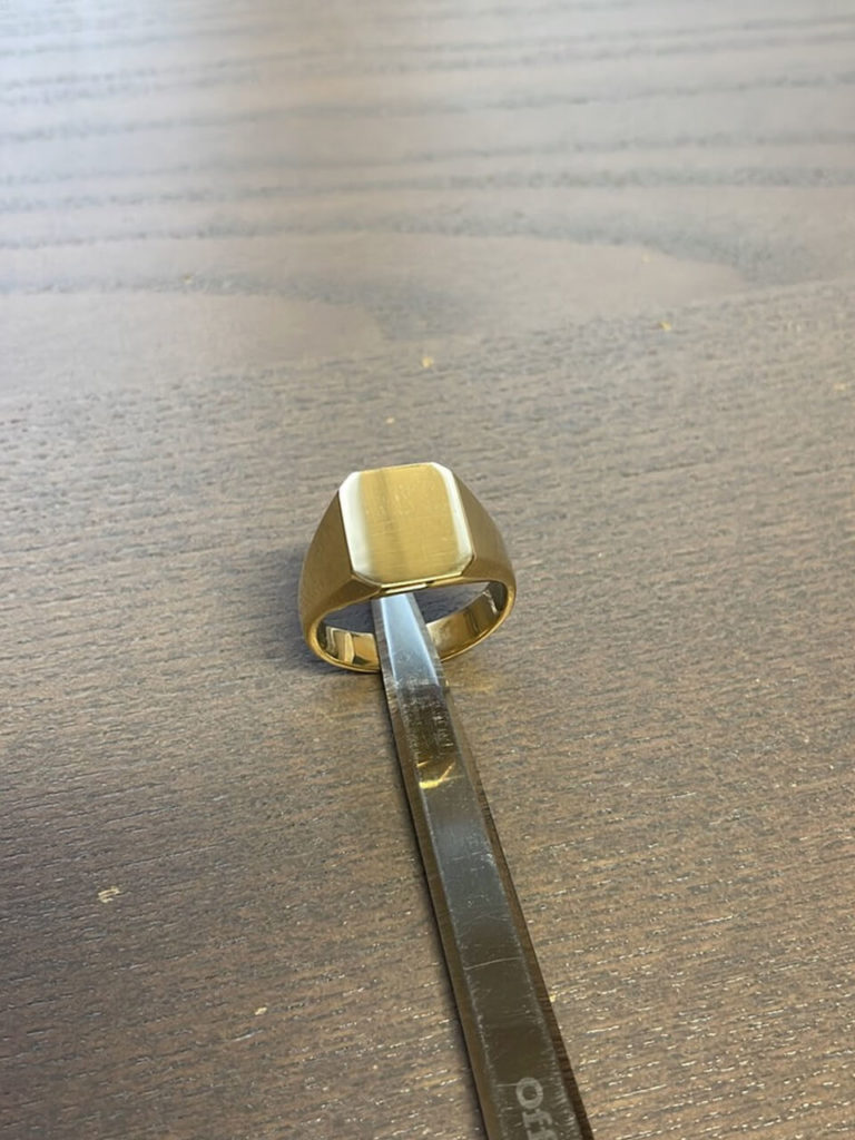 Ein eleganter, goldener Siegelring mit glatter Oberfläche, der von einer Pinzette gehalten wird.