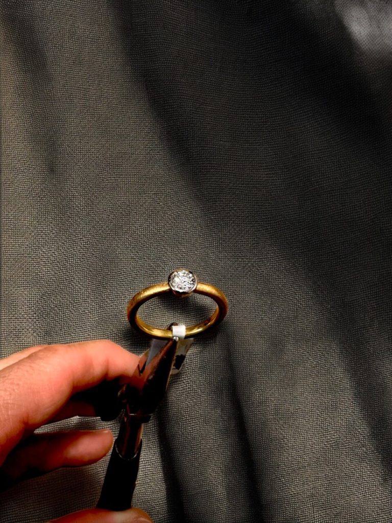 Ein eleganter Goldring mit funkelndem Diamanten wird bei Goldwerk Berlin-Lankwitz fachmännisch mit einer Zange für eine Reparatur gehalten.