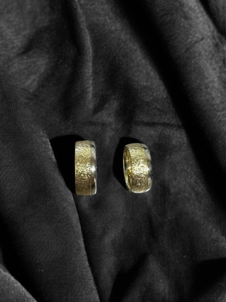 Zwei elegante Eheringe mit strukturierter, goldener Oberfläche und glatten silberfarbenen Rändern auf einem dunklen, weichen Stoffhintergrund.