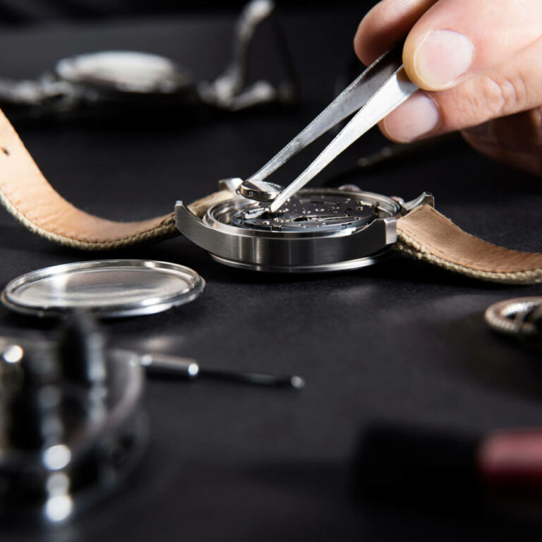 Ein Uhrmacher setzt mit einer Pinzette eine kleine Batterie in eine Armbanduhr ein. Professioneller Service für Uhren und Schmuck in Berlin-Lankwitz.